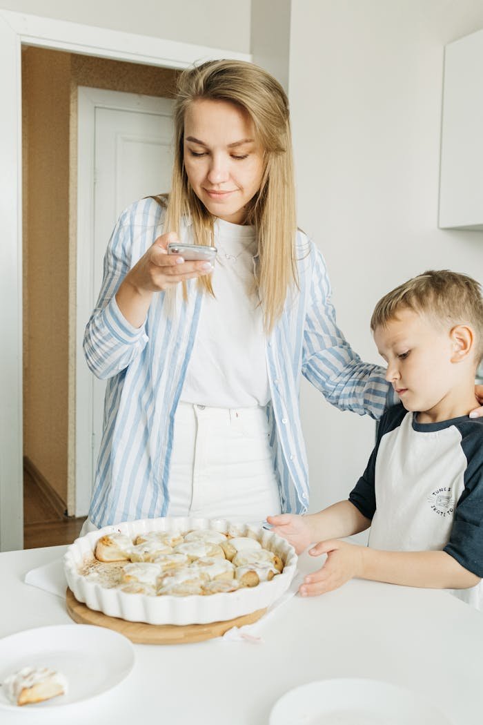 gallery-02 A mother taking a photo of freshly baked cinnamon rolls with her son in a cozy kitchen setting.